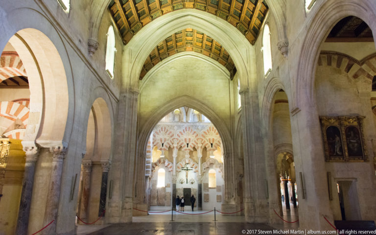 Tall pale cathedral bay opening toward a distant group of red-and-light striped arches inside the Mezquita.
