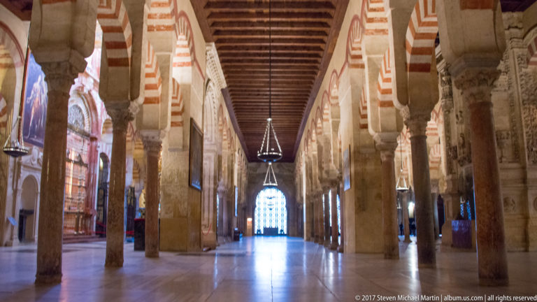Long timber-roofed Mezquita aisle with stone columns, striped arches, hanging lamps, and a bright opening at the end.
