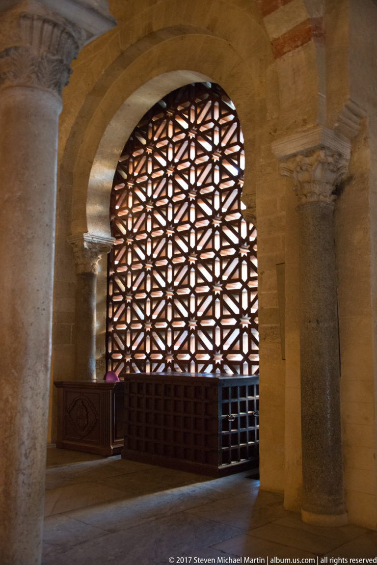 Dark geometric wooden lattice screen glowing within a broad stone arch beside massive masonry supports.