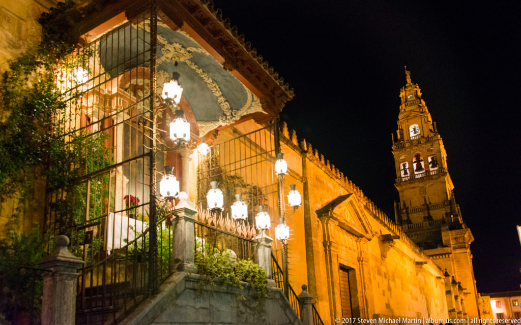 Illuminated exterior shrine beside the Mezquita wall, with the bell tower rising in the distance at night.
