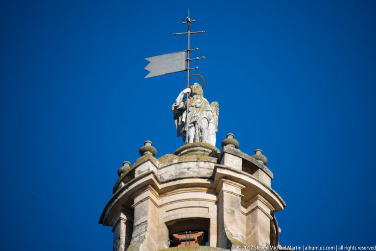 Close view of the Mezquita bell tower crown with a sculptural figure, metal weather vane, and corner finials.