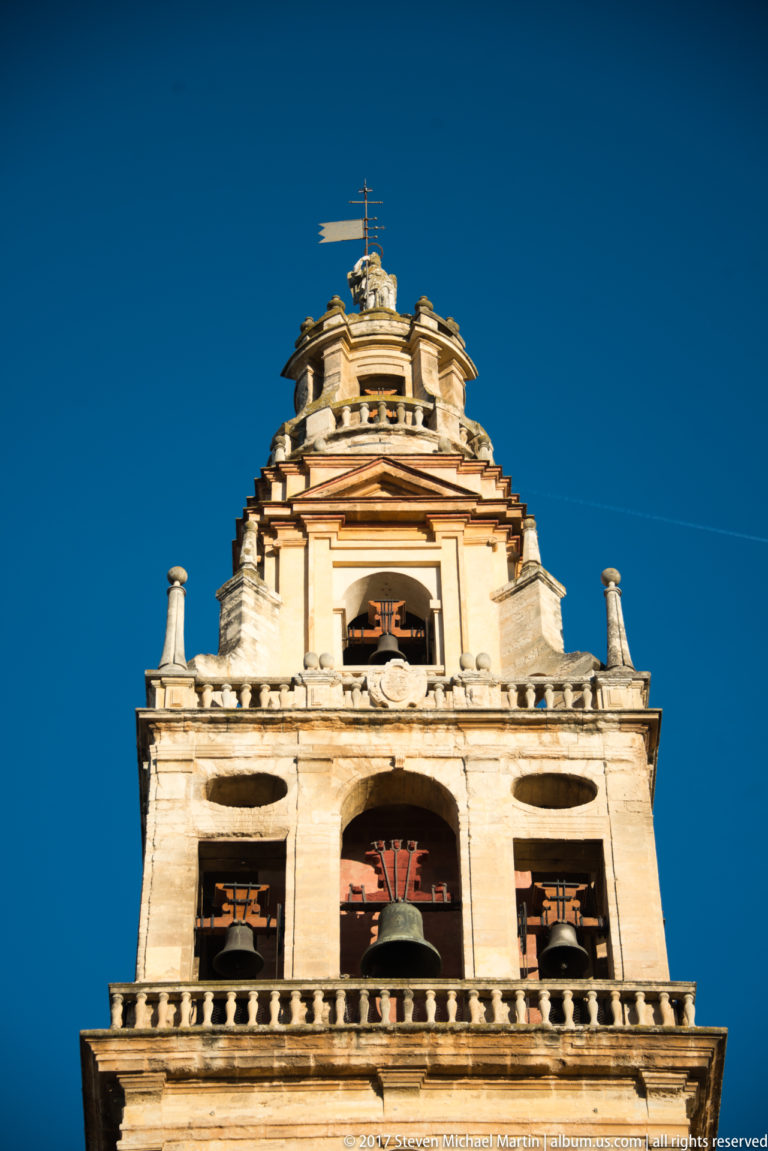Close frontal view of the Mezquita bell tower’s upper stages, with bells, balustrades, finials, and sculptural crown.