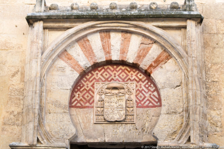 Sealed horseshoe arch with red-and-pale voussoirs, geometric decoration, and a carved heraldic plaque on the Mezquita exterior.