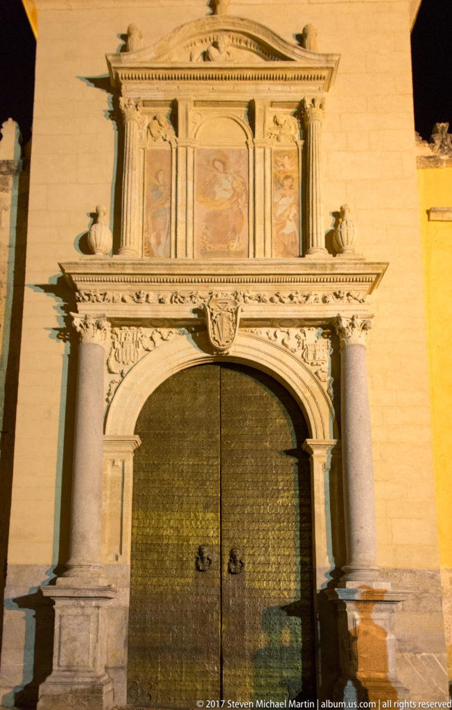 Classical stone portal with paired columns, carved ornament, heraldic shield, and large dark metal doors.