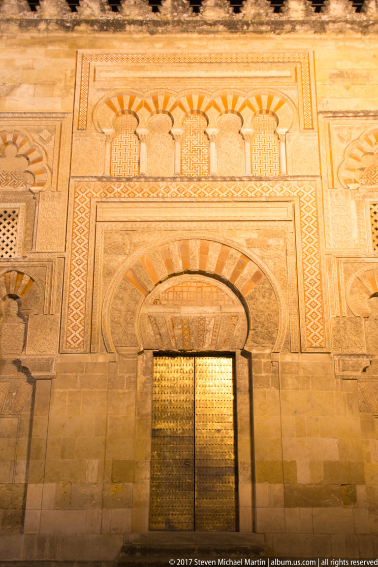 Ornately decorated horseshoe-arched doorway with striped voussoirs, geometric panels, and metal doors on the Mezquita exterior.