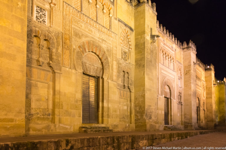 Oblique night view of several decorated horseshoe-arched portals receding along the Mezquita’s exterior wall.