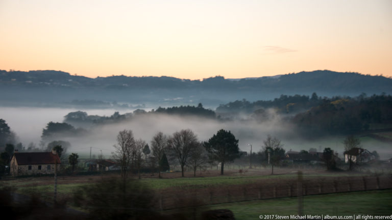 Morning fog over fields, houses, and wooded hills beneath a pale sunrise.