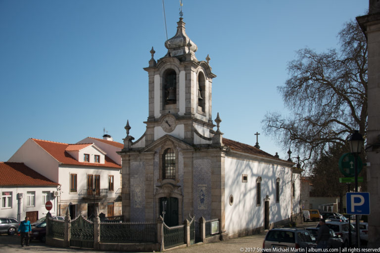 Stone church with arched doorway, clock, ornate open bell tower, and white side walls.