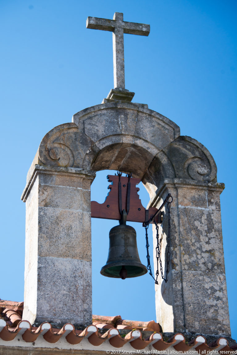 Bronze bell hanging from a red timber yoke in a granite bell-cote topped by a cross.
