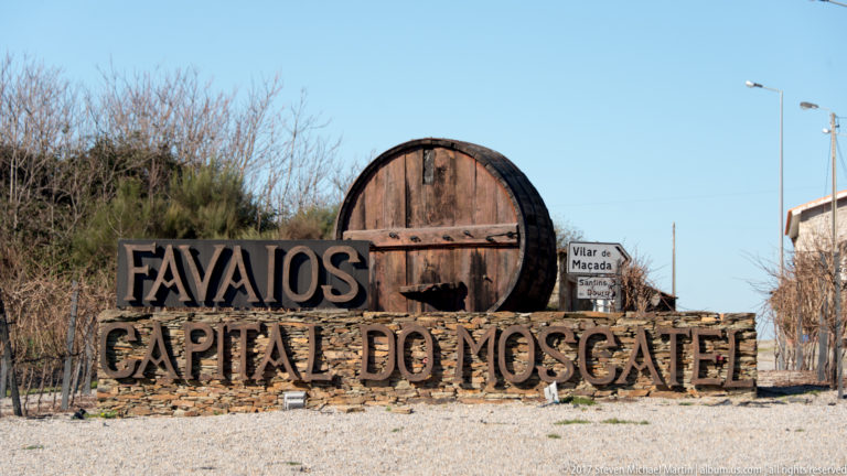 Roadside sign reading “Favaios Capital do Moscatel” beside a large wooden barrel end.