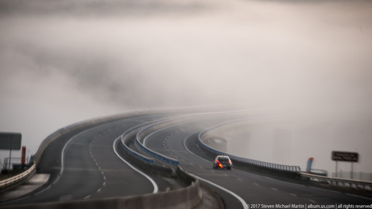 Curving divided highway with one car disappearing into dense fog.