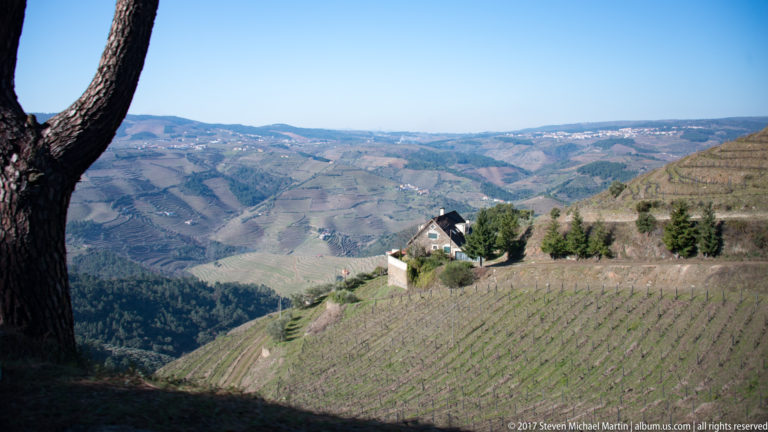 Steep vineyard below a hillside house overlooking layered cultivated mountains and valleys.