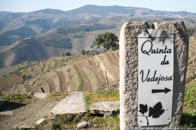 Stone sign reading “Quinta da Vedejosa” with an arrow and grape-leaf motifs before vineyard hills.