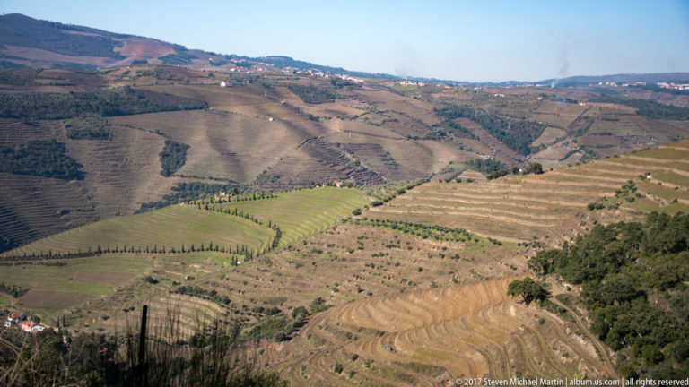 Wide cultivated mountain landscape with vineyards, orchards, and patterned fields across rolling slopes.
