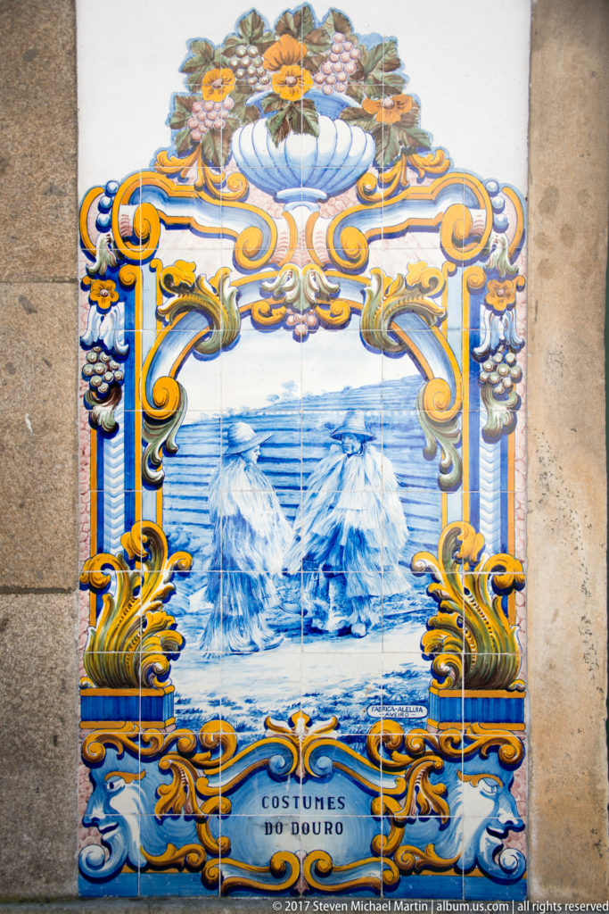 Small azulejo panel labeled Costumes do Douro, showing two conical haystacks beneath an ornate floral and scroll frame.
