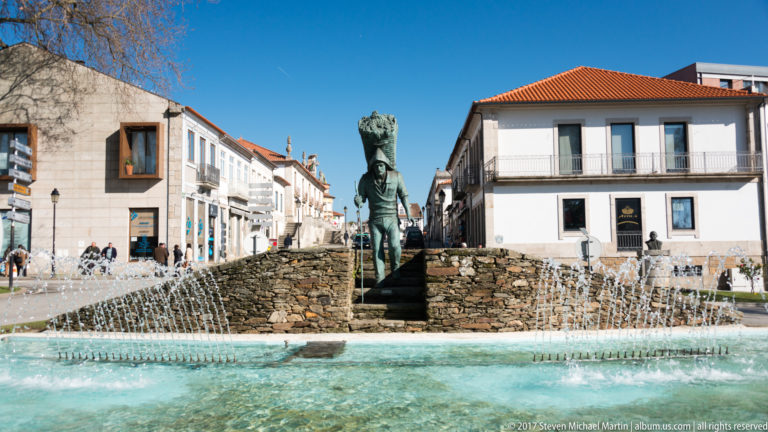 Bronze figure carrying a large basket in a circular fountain surrounded by white buildings.