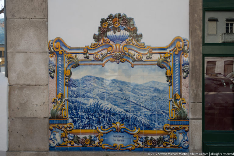 Panorama do Pinhão azulejo dominated by layered cultivated hills and vineyard rows within an ornate yellow and blue border.