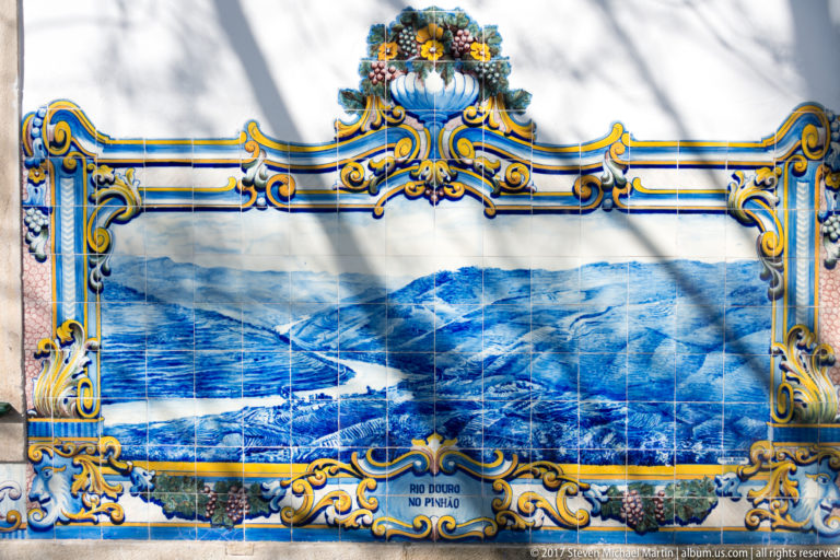 Sunlit azulejo panel labeled Rio Douro no Pinhão, with river and mountain landscape crossed by tree shadows.
