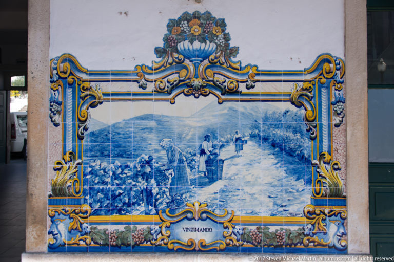 Azulejo panel labeled Vindimando, with vineyard workers walking along a path between grapevines and hills.