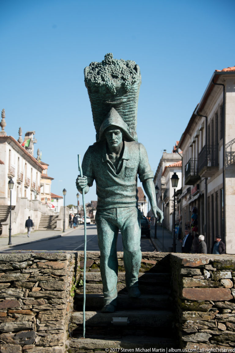 Close view of a bronze worker carrying a large basket and holding a long staff.