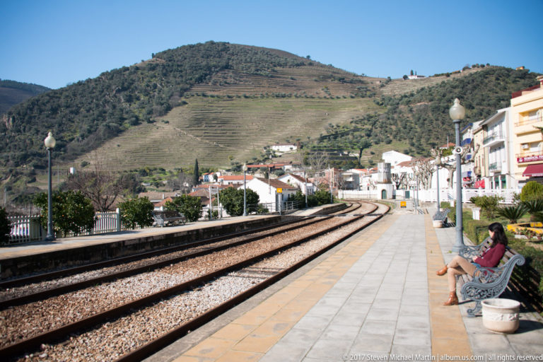 Curving railway tracks beside Pinhão station platform, with a seated passenger and terraced vineyard hills rising behind the town.