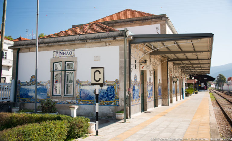 Pinhão railway station building with tiled roof, platform canopy, PINHÃO sign, and blue-and-white azulejo panels along the façade.