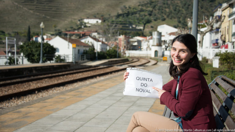 Smiling woman seated on Pinhão station platform holding a Quinta do Noval sign, with curving tracks and hillside buildings behind.