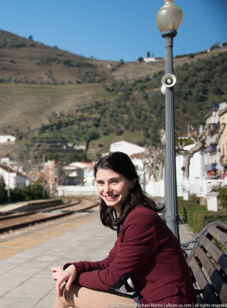 Woman seated on a wooden bench at Pinhão station, with railway tracks, platform lamps, town buildings, and vineyard hills behind.