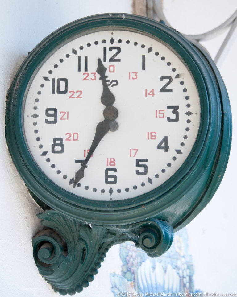 Close view of a round Pinhão station clock with black numerals, smaller red 24-hour numbers, and ornate green metal casing.