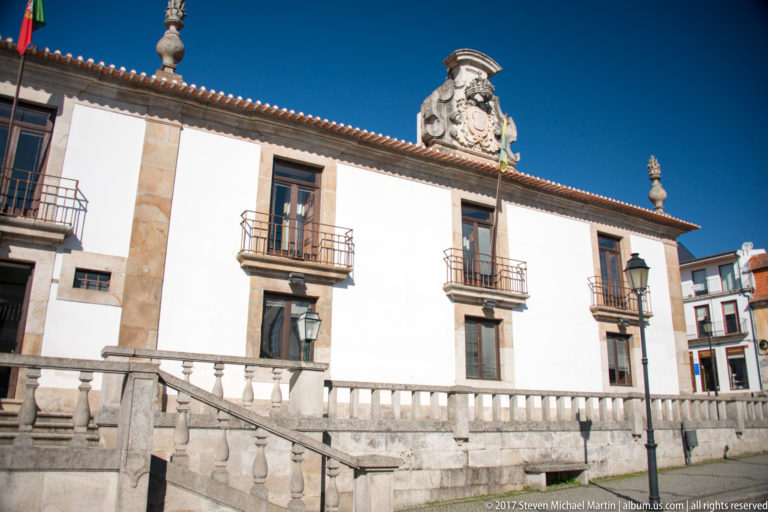 White civic-looking building with stone trim, iron balconies, balustrade, and a large heraldic crest.