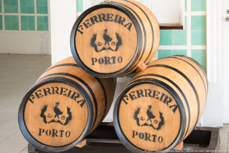 Three stacked wooden barrels branded “FERREIRA PORTO,” displayed indoors against pale walls and turquoise tile.