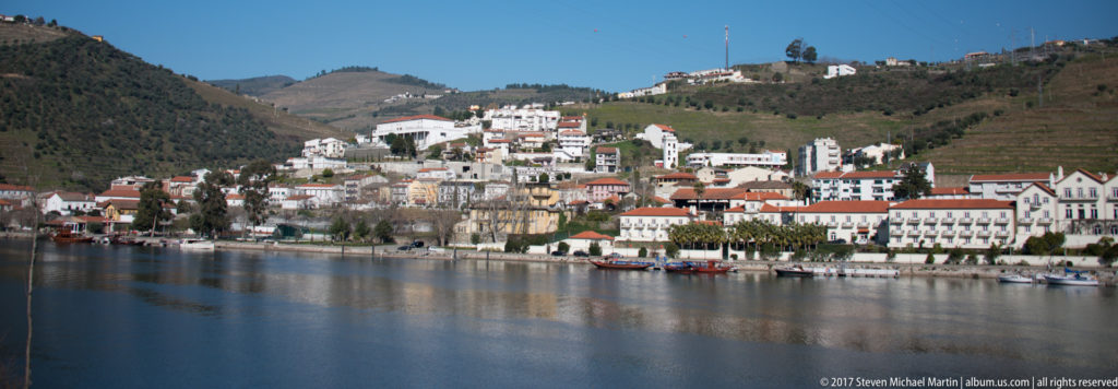Pinhão’s white and red-roofed riverfront reflected in calm Douro water, with terraced hills rising behind.