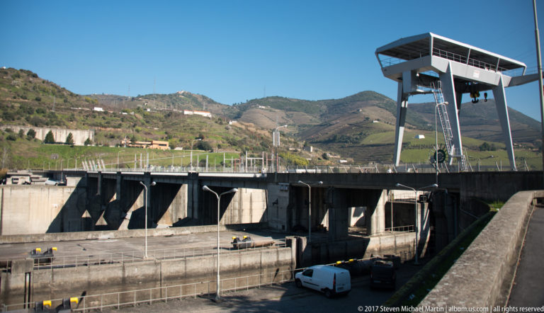 Concrete navigation lock with gates, service platforms, and an elevated metal lifting structure against terraced hills.