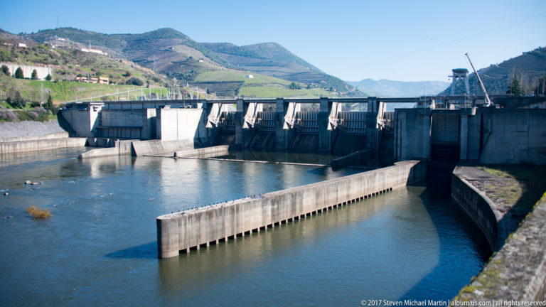 Concrete dam with gated spillway and divided navigation channel across the Douro, backed by vineyard-covered mountains.