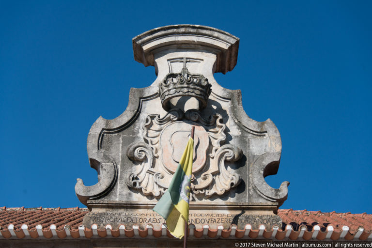 Carved heraldic crest with shield and crown above a tiled roof, partly crossed by a flag.