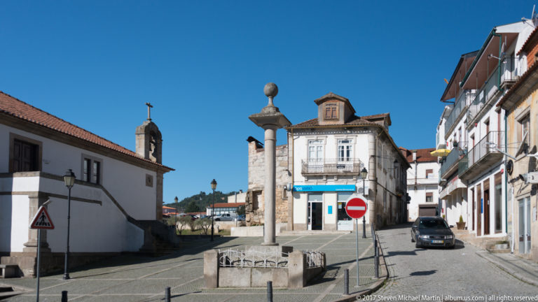 Stone pillory on a raised square beside a narrow street of white and stone buildings.
