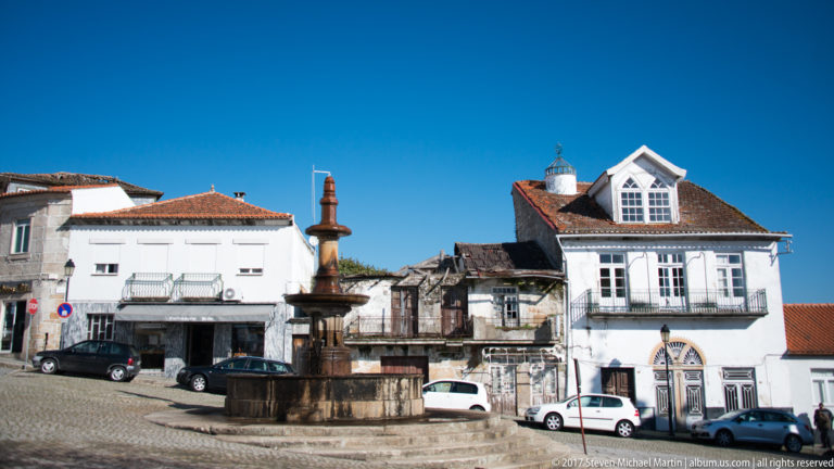 Tiered stone fountain or monument in a cobbled square surrounded by whitewashed and weathered houses.