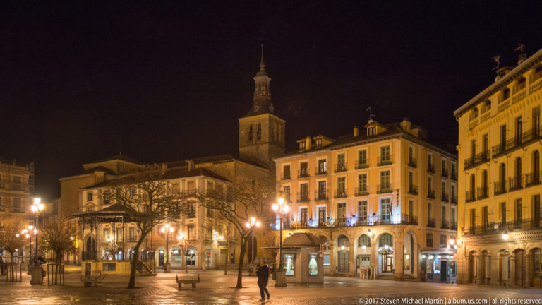 Night view across Plaza Mayor with arcaded buildings, lit shopfronts, a church tower, and a few pedestrians crossing the square.