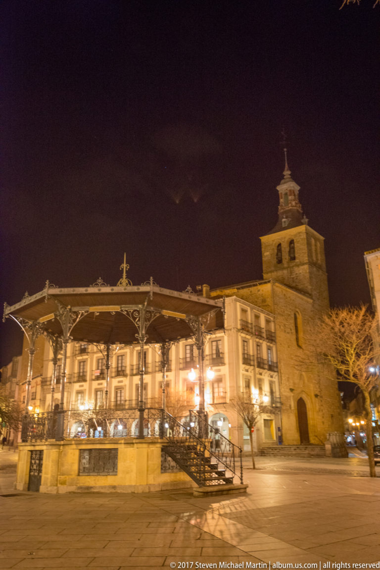 Night view of Plaza Mayor with a large covered bandstand in the foreground and a stone church tower rising to the right.