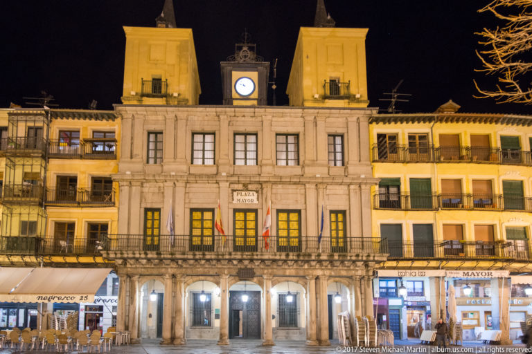Symmetrical civic building on Plaza Mayor with a central clock, twin corner towers, long balcony, and ground-floor arcade.