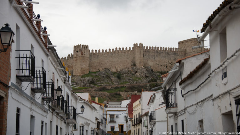 Whitewashed Andalusian street leading uphill toward a crenellated stone castle on a rocky ridge.