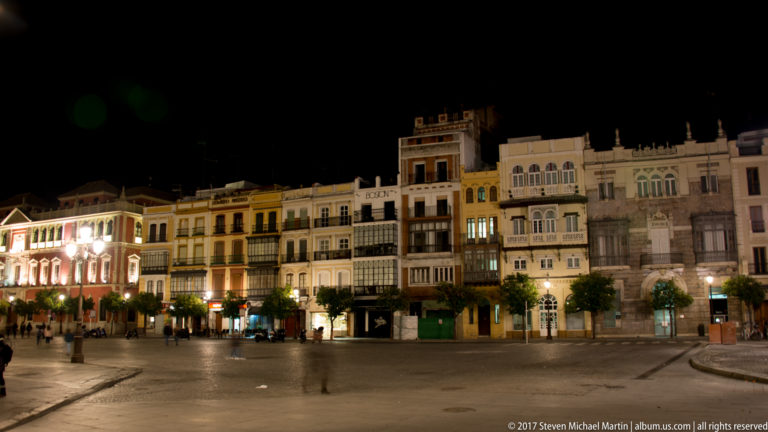 Illuminated historic façades with balconies lining a broad Seville square at night.