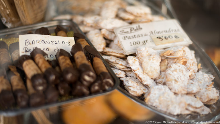 Tray of dark rolled barquillos beside pale almond pastries with handwritten price labels in a pastry shop.