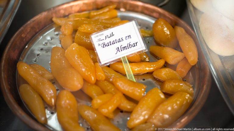 Glossy orange batatas en almíbar sit in a round metal bowl beneath a handwritten label.