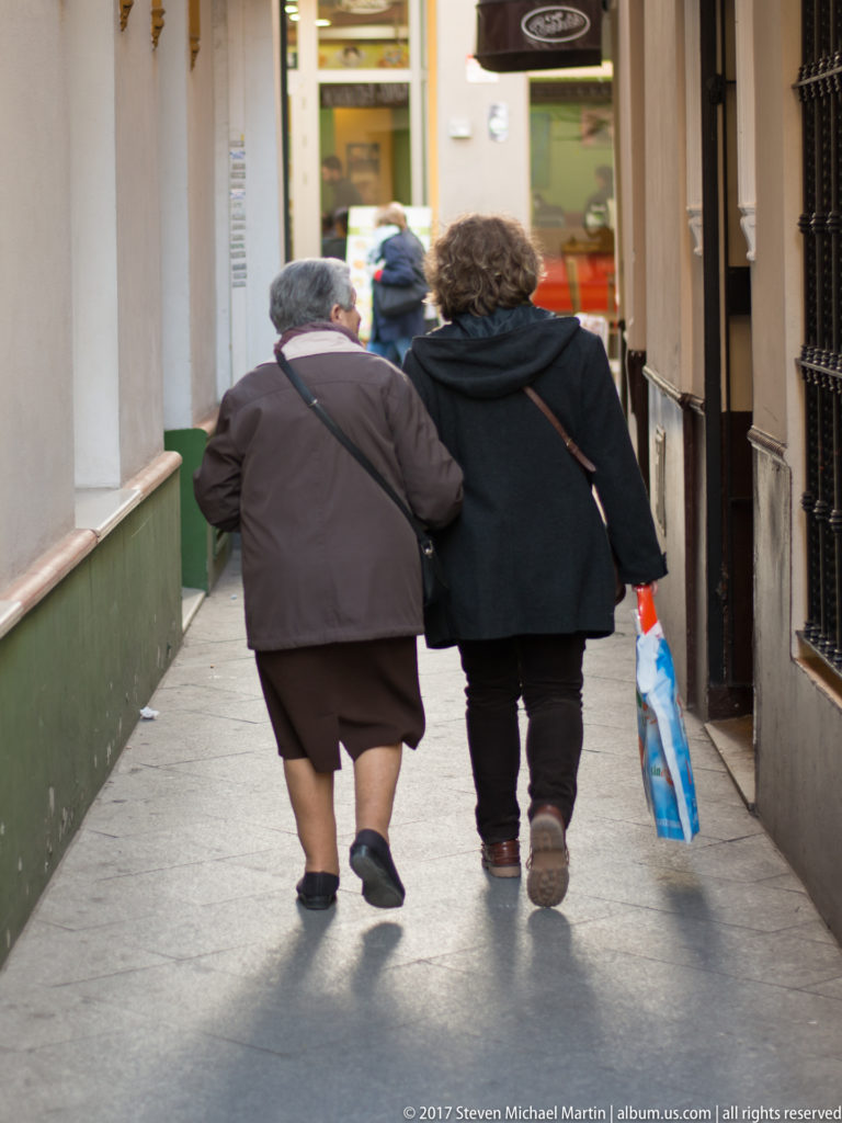 Two women walk arm in arm away from the camera along a narrow Seville sidewalk, one carrying a blue bag.