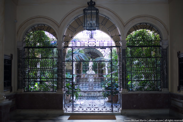 Three arched wrought-iron gates and a hanging lantern frame a bright courtyard filled with palms and greenery.