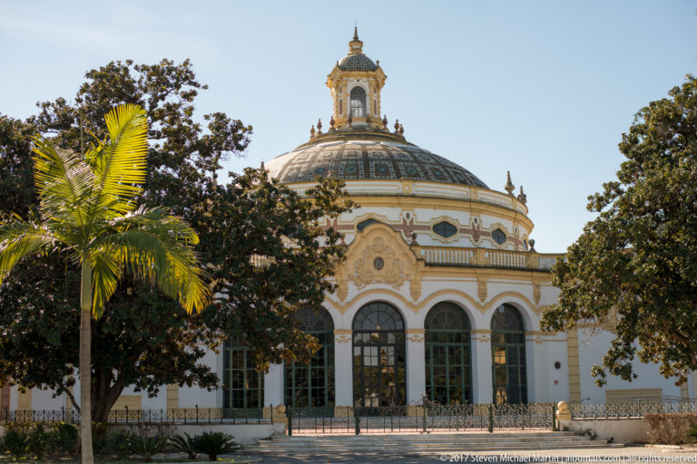 Ornate domed pavilion with arched windows and cupola stands behind palms and mature trees in a Seville park.