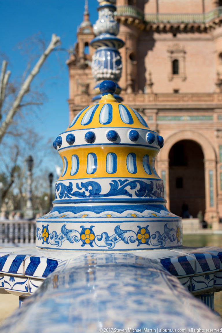 Blue-and-yellow glazed ceramic finial in sharp focus with the brick architecture of Plaza de España blurred behind.