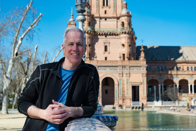 Man poses beside the canal at Plaza de España with a brick tower and arcaded façade behind him.