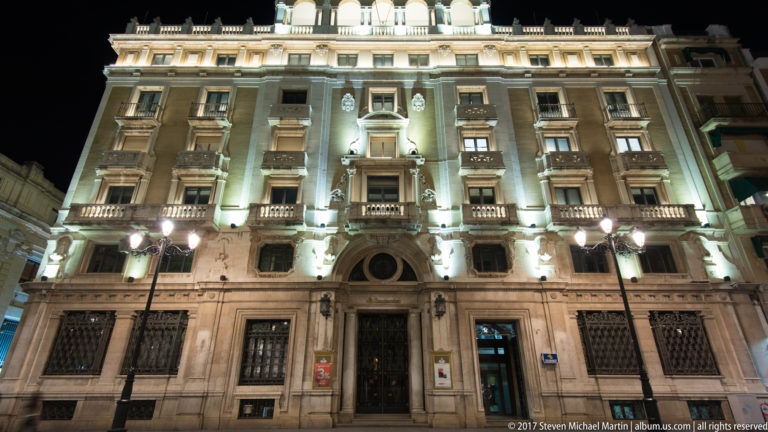 Illuminated ornate stone façade with arched entrance, stacked balconies, sculptural decoration, and a high cornice.